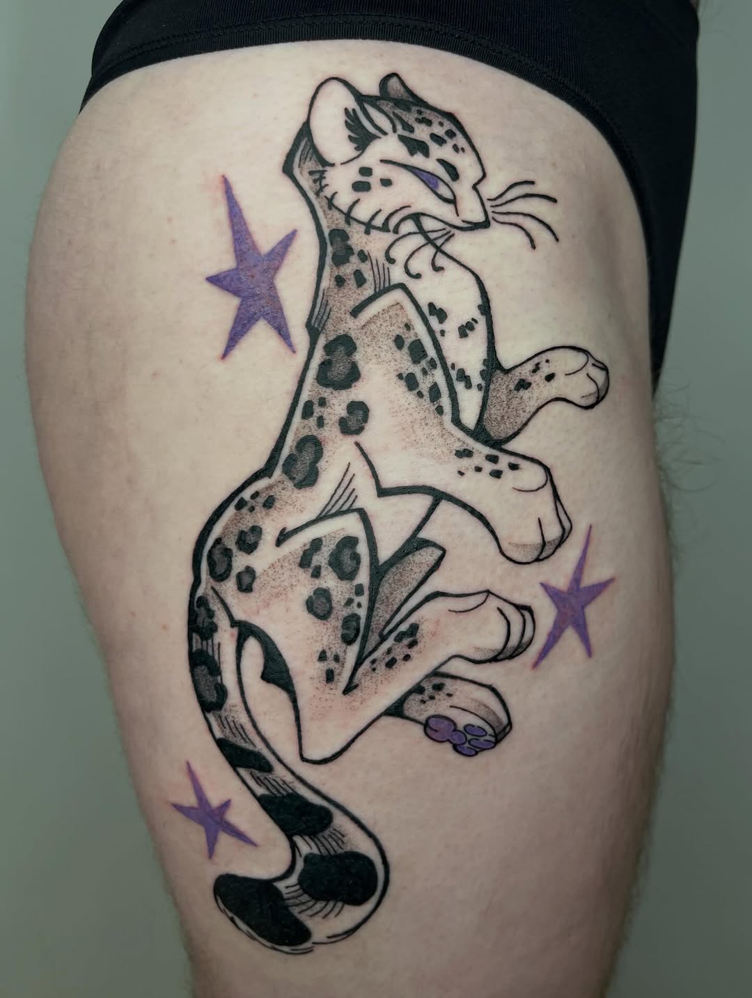 Spotted ocelot or wild cat tattoo on thigh with purple stars, featuring black ink outlines, shading, and tan/brown toning on pale skin.
