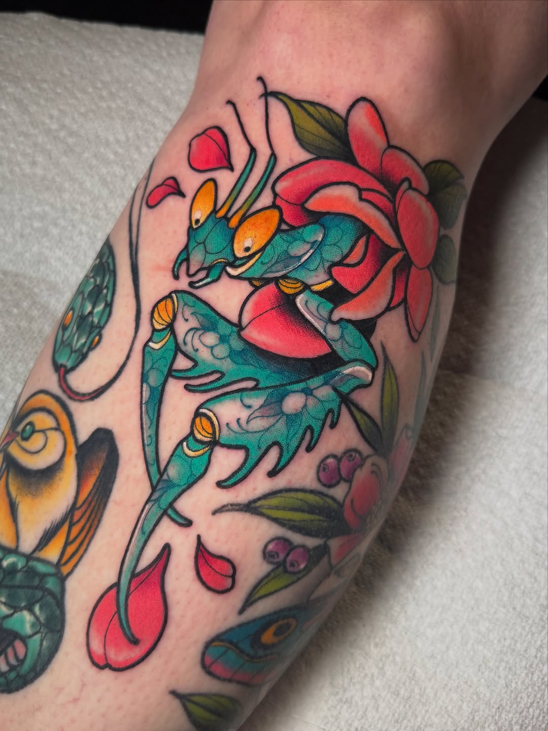 Colorful tattoo on a calf featuring three teal insects with orange eyes on red flowers, green leaves, and purple berries.