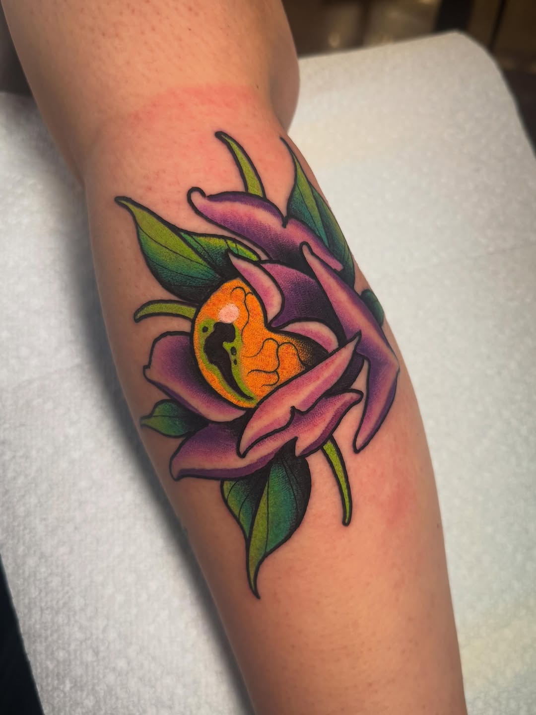 Colorful flower tattoo on the forearm featuring pink petals, orange center with green details, and dark outlines against skin.