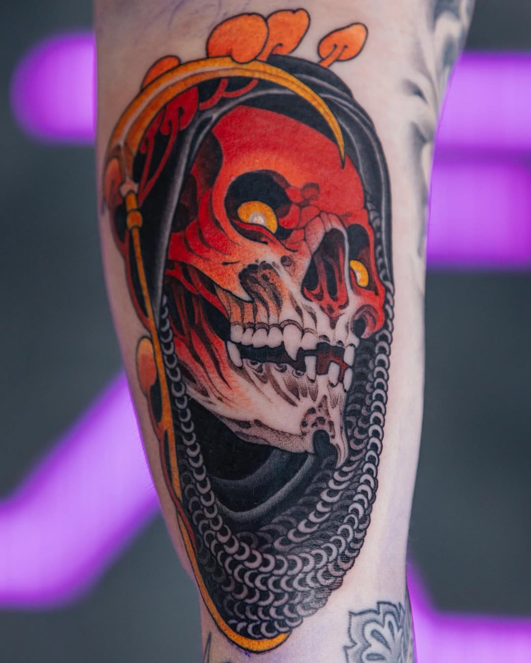 Red and orange skull with golden eyes and black decorative patterns, framed by an orange oval border on the leg.