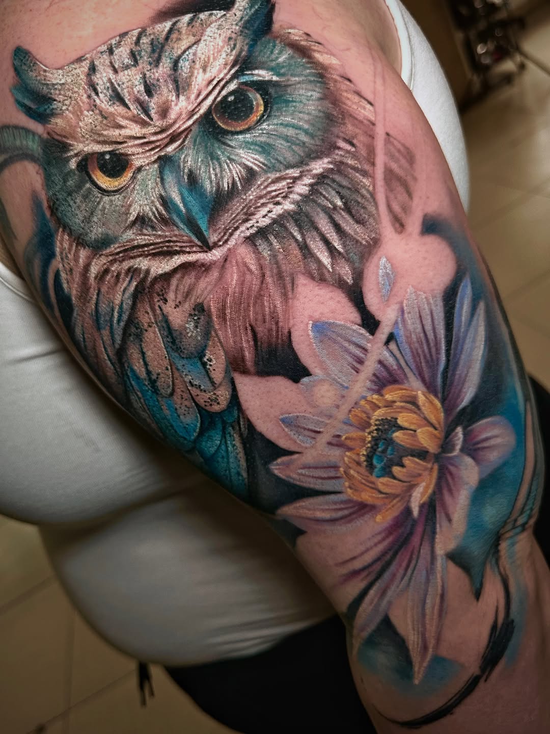 Detailed owl tattoo with golden eyes and feathered plumage on the shoulder, accompanied by purple and yellow flowers on the upper arm.
