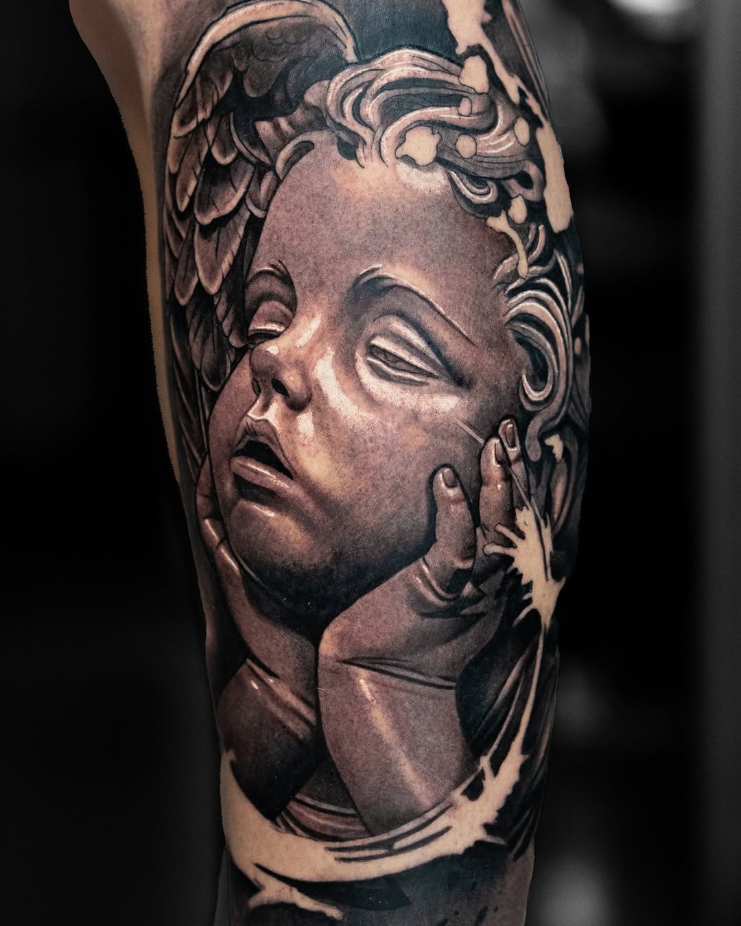 Detailed portrait tattoo of a classical male head with curled hair and closed eyes on the upper leg, rendered in black and grey ink.