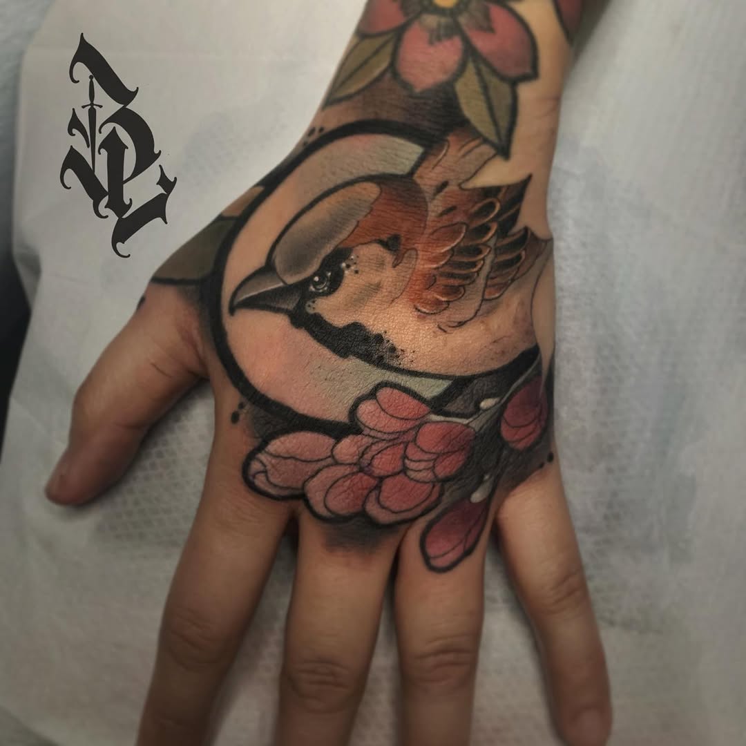Hand tattoo featuring a winged bird's head in profile with red and tan coloring, flowers above, and a decorative symbol on the wrist.