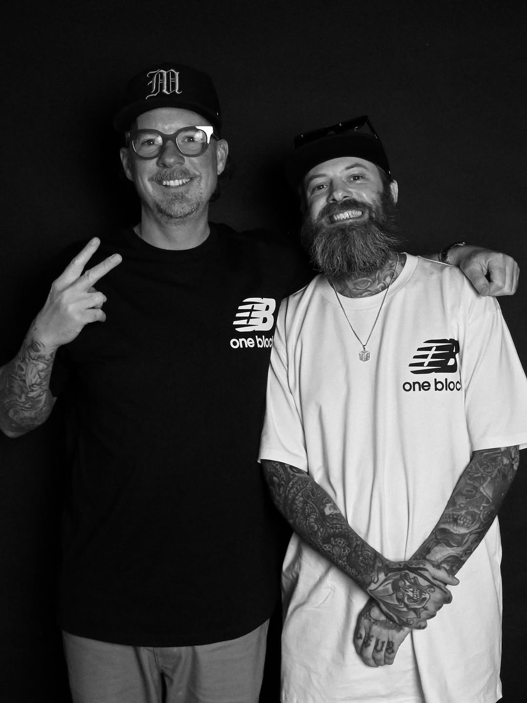 Two men posing together, the man on the right displays extensive black tattoos covering both arms and hands against his white shirt.