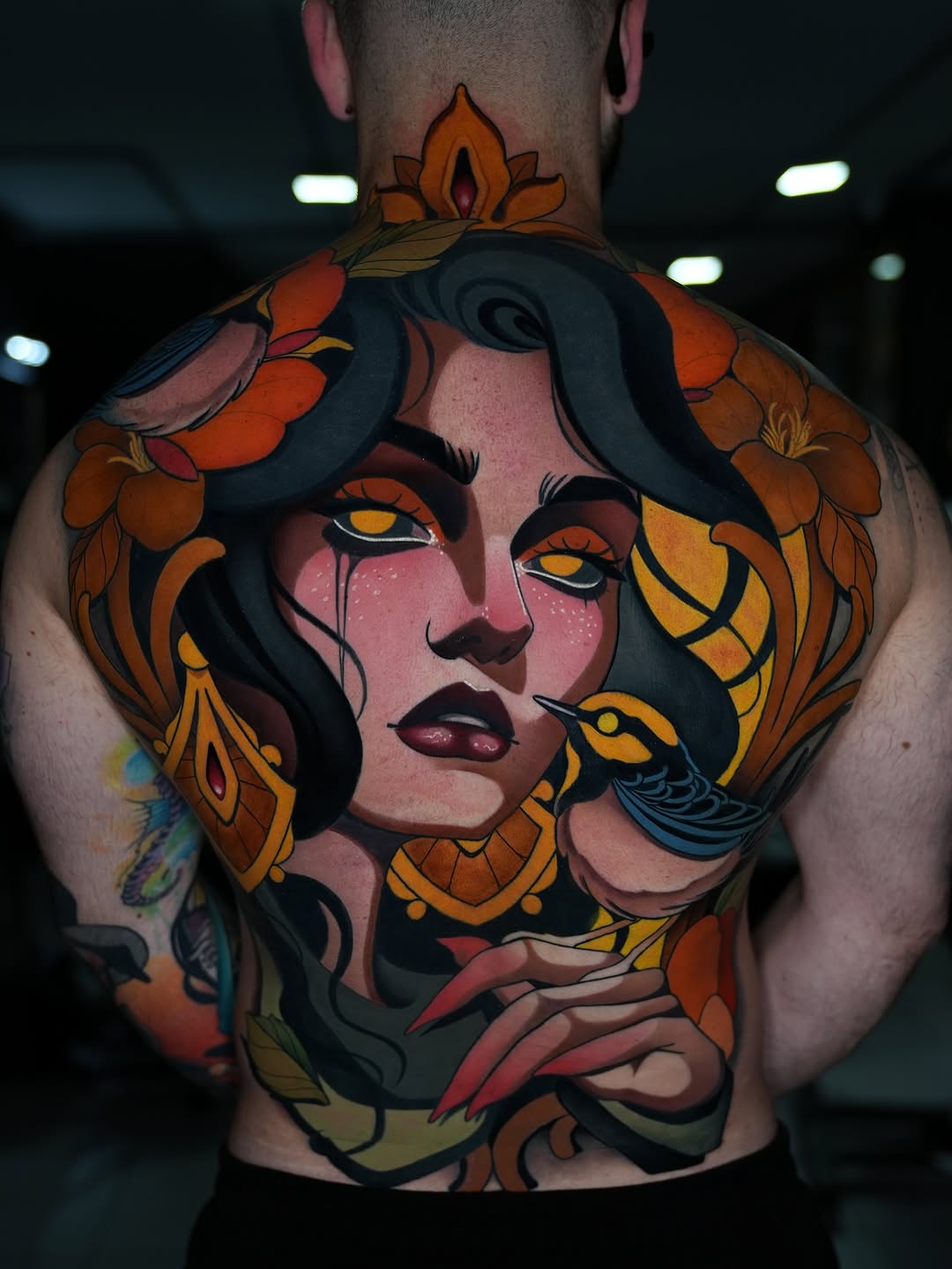 Large portrait tattoo on upper back and shoulders featuring a woman's face with black hair, golden-yellow eyes, and pink skin surrounded by orange and red flowers.