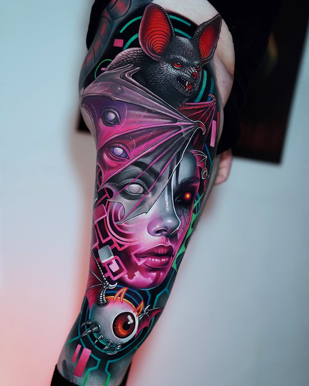Colorful full-sleeve tattoo featuring a bat-eared creature, human face, geometric patterns, and glowing eyes in pink, magenta, teal, and black on the arm.