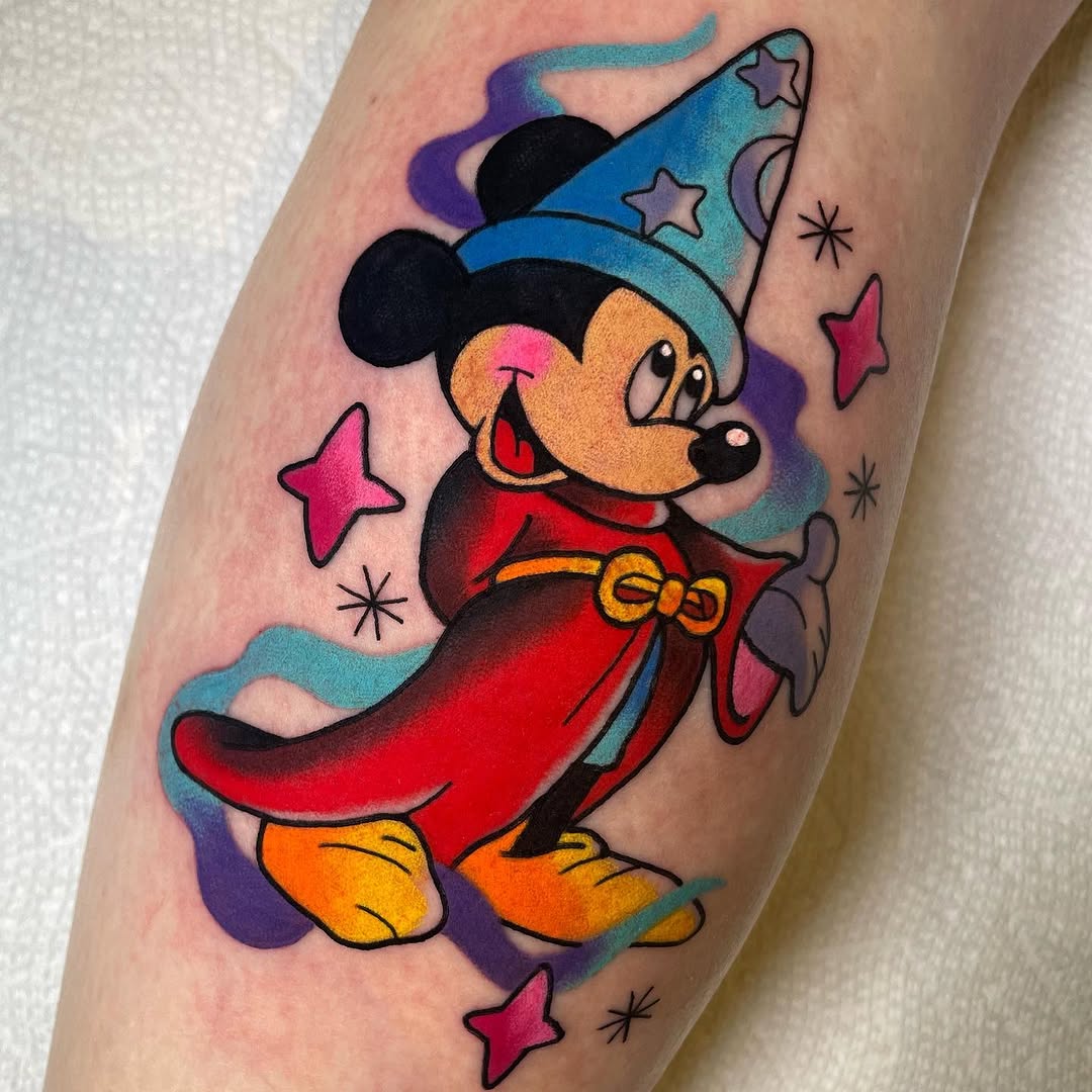 Colorful Mickey Mouse tattoo wearing a blue wizard hat and red cape, surrounded by pink stars and sparkles on the upper arm.