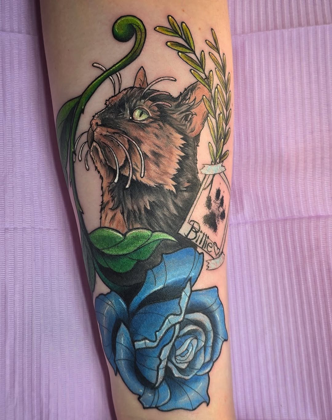 Colorful cat tattoo on forearm featuring a tabby cat with green vine and leaf elements, blue roses, and text reading "Blurred," displayed on pale skin.