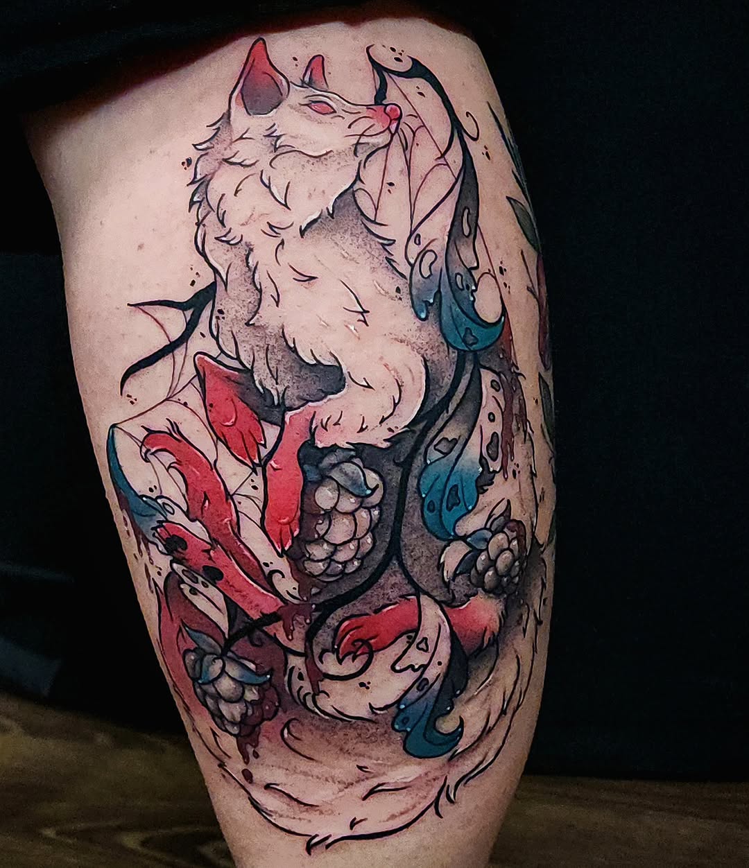 Colorful leg tattoo featuring two stylized wolves with red and teal accents, ornamental vine details, and decorative elements on the calf.