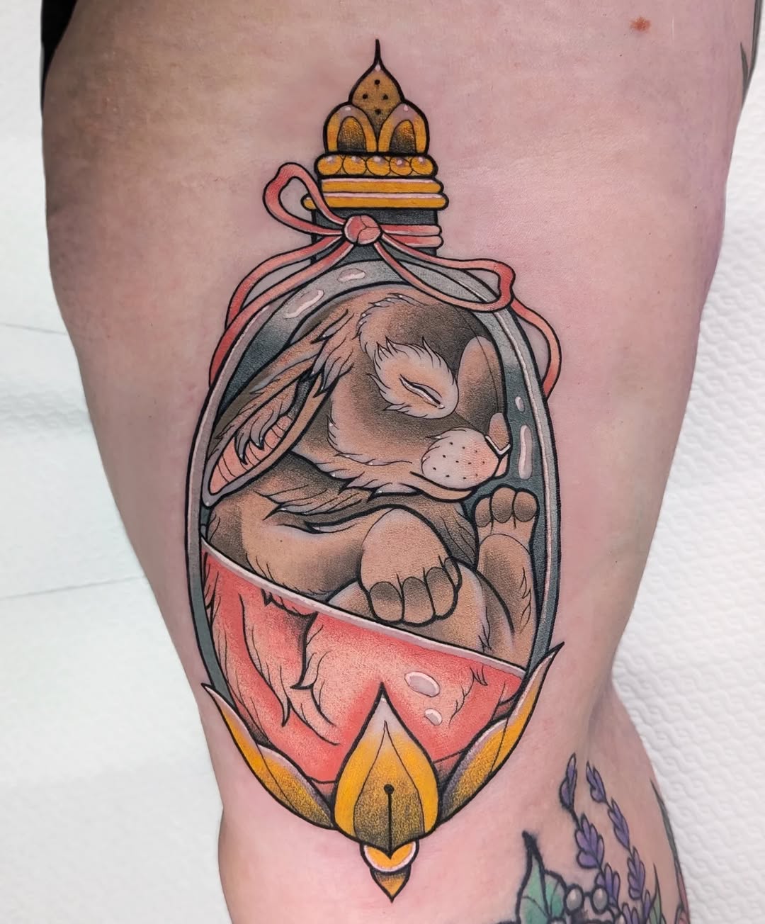 Colored tattoo on thigh depicting a sleeping animal in ornate frame with red ribbon bow, golden crown top, and lotus flower bottom.