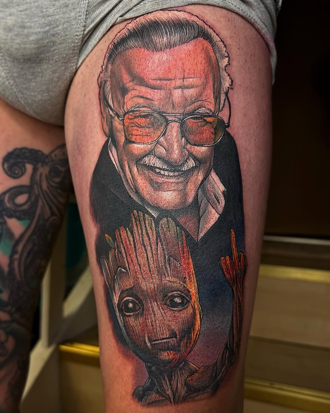 Portrait tattoo on thigh featuring a smiling man with glasses above a wooden alien head, rendered in red, black, and skin tones.