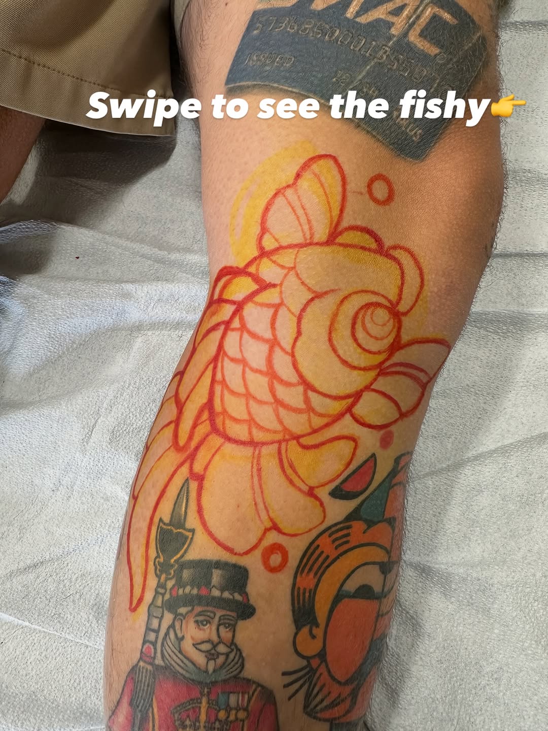 Colorful koi fish tattoo in red, yellow, and orange on the leg, positioned above other existing tattoos featuring a figure in a hat.