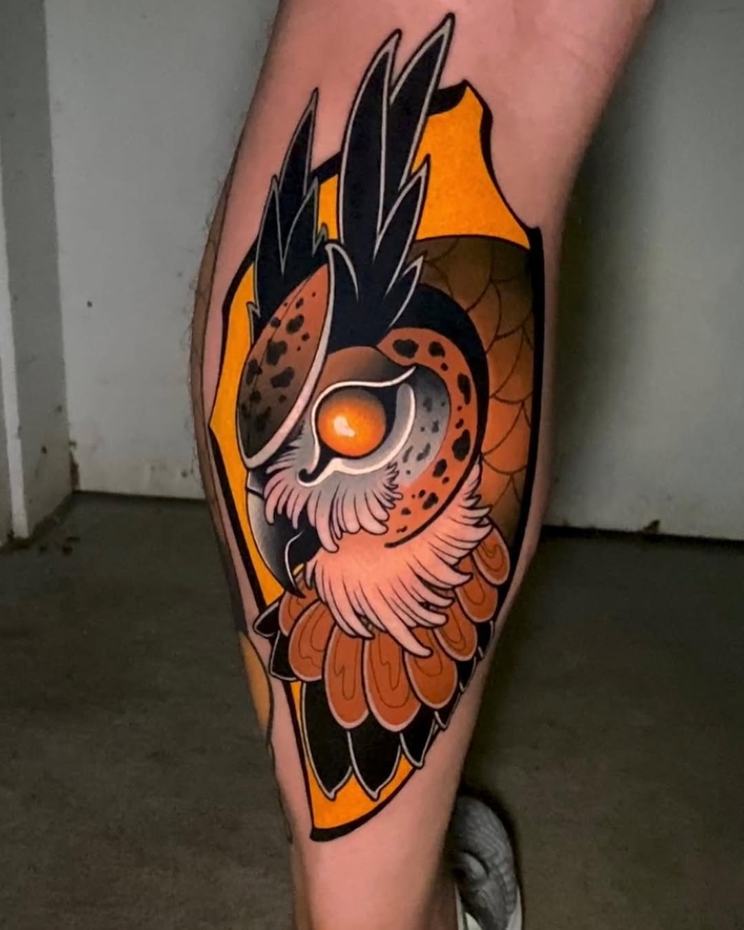 Orange and black owl tattoo on the calf with sharp pointed feathers, large glowing orange eyes, and detailed wing patterns.