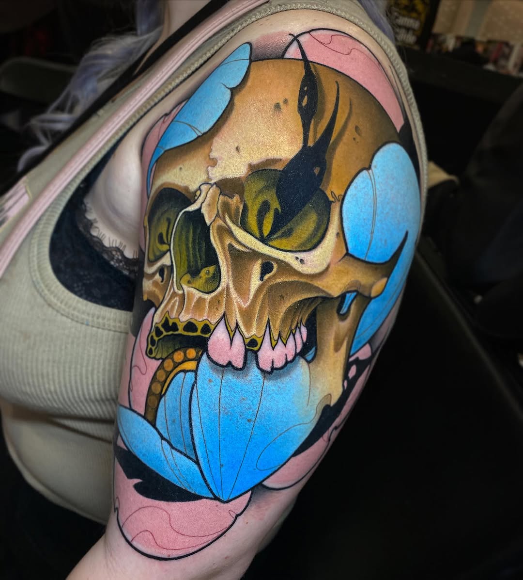 Colorful skull tattoo on upper arm featuring blue petals, pink background, golden skull with green eyes and yellow teeth details.