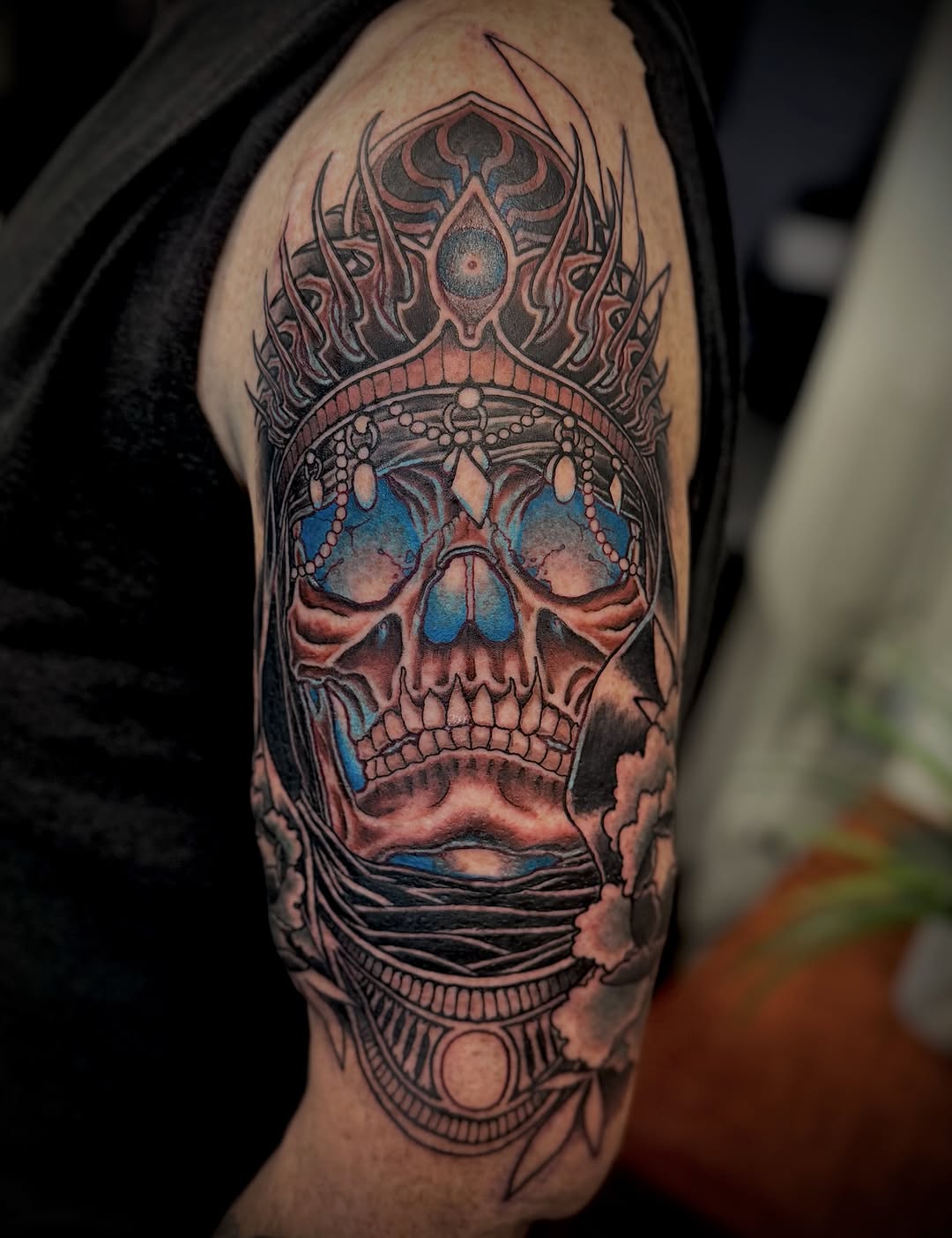 Ornate skull with blue eye sockets and jeweled crown on upper arm, featuring intricate decorative patterns in red, blue, and grey ink.