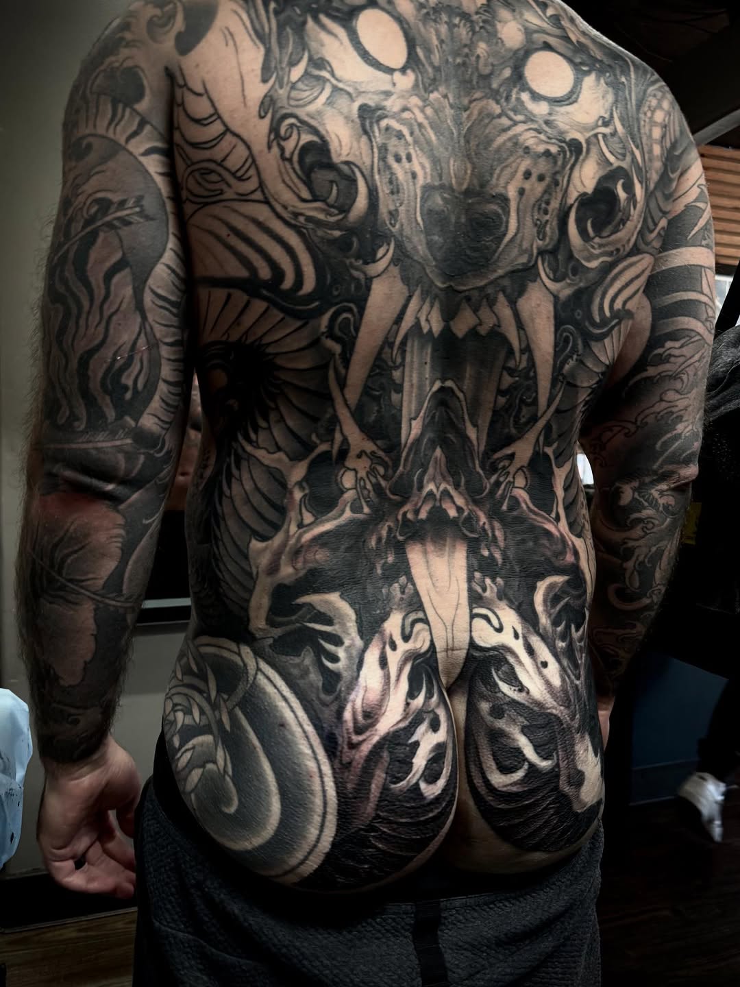 Back and torso tattoo featuring ornate skulls, flowing scrollwork, and decorative elements in black and gray ink covering the shoulders, spine, and lower back.
