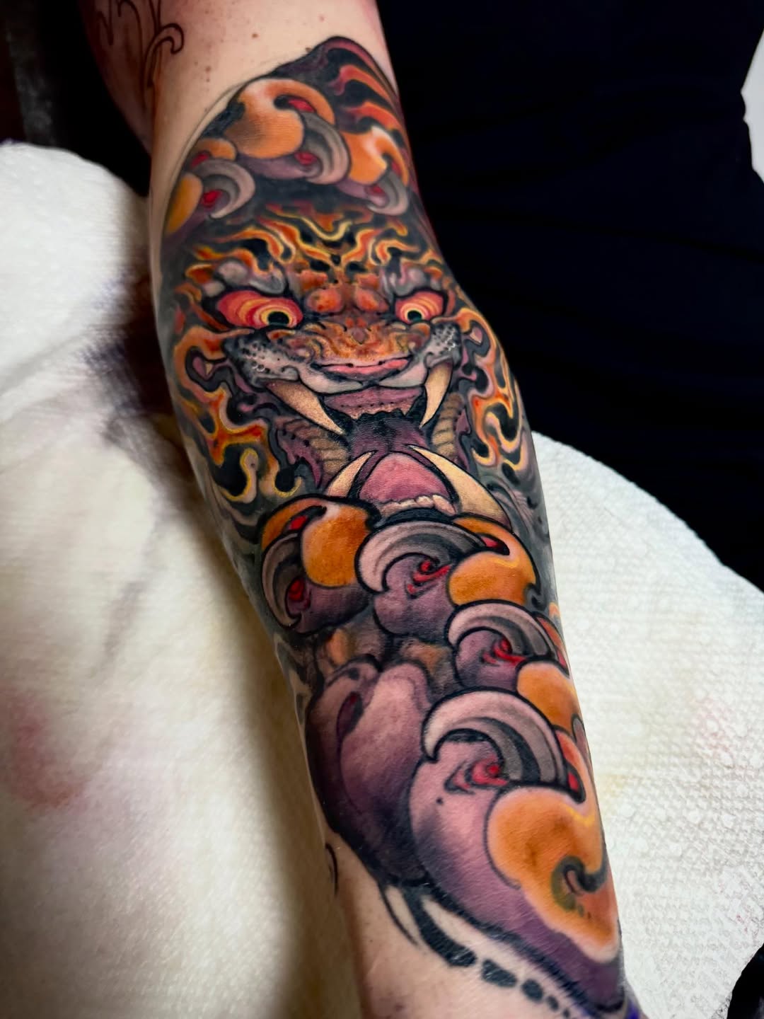 Colorful oni mask tattoo on forearm featuring orange flames, purple clouds, and red eyes, with decorative scrollwork below in orange and mauve tones.