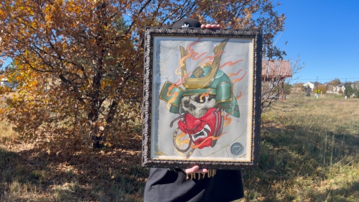 Tattoo design featuring a red oni mask with green samurai armor and gold accents, held up outdoors against autumn foliage.