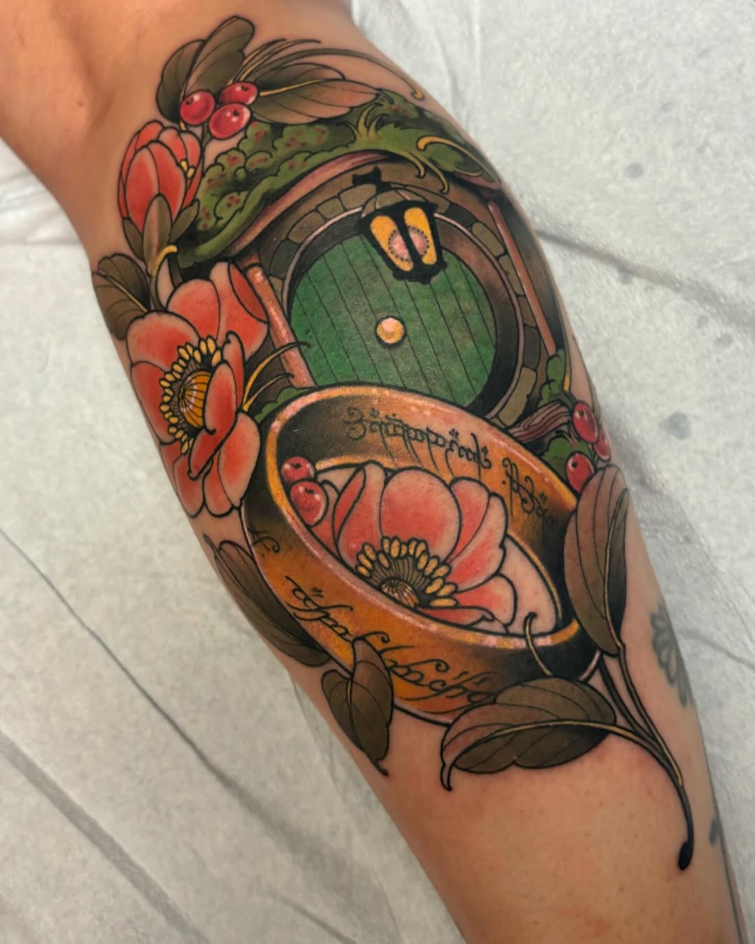 Colorful leg tattoo featuring a hobbit hole door with glowing eyes, surrounded by red flowers, berries, and decorative foliage in warm earth tones.
