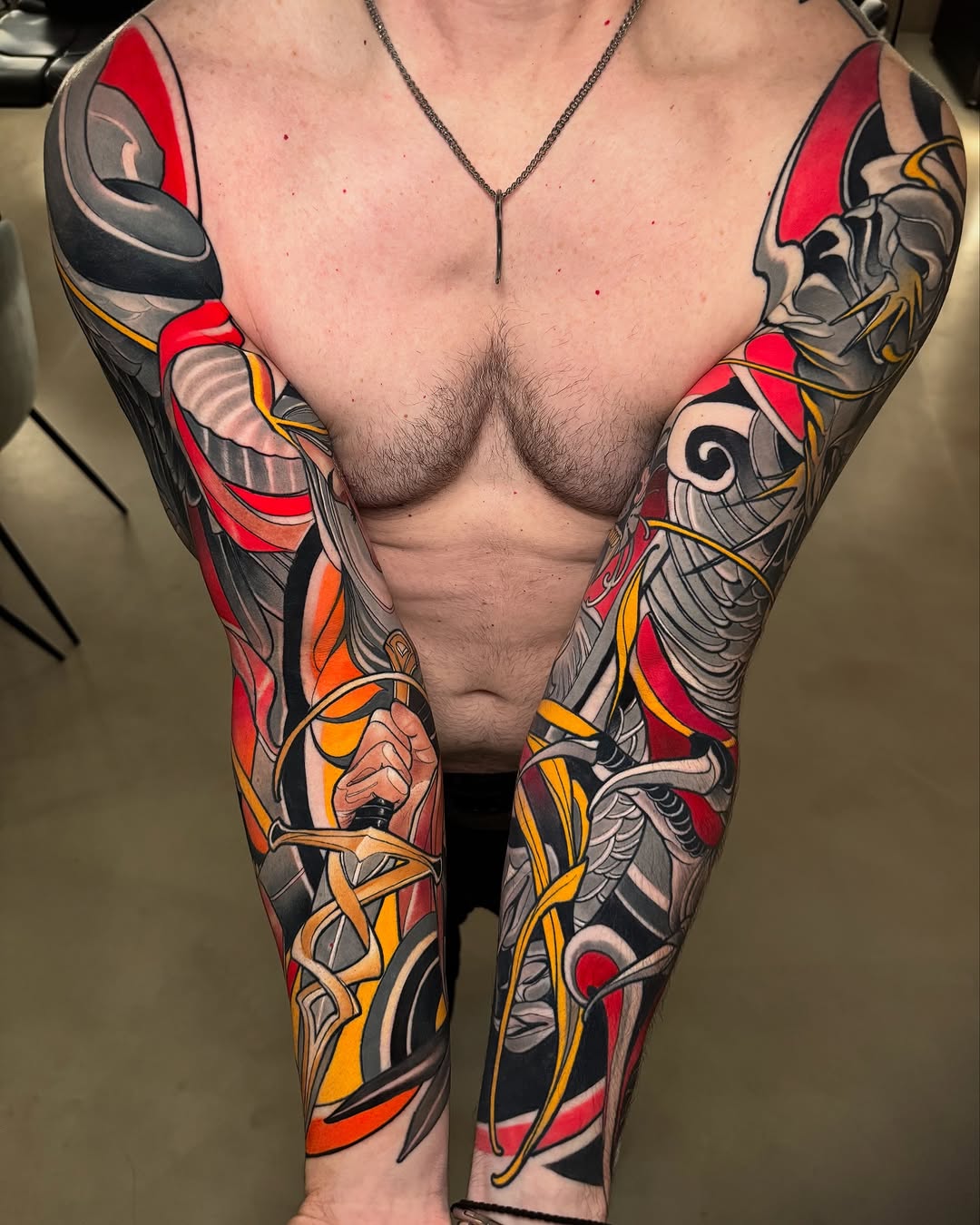 Full-sleeve arm tattoos featuring red, orange, black, and gold geometric and abstract designs covering both shoulders, biceps, and forearms.