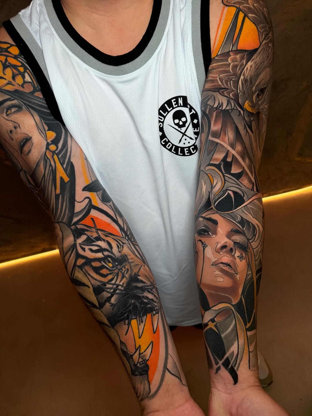 Full-sleeve arm tattoos featuring portraits of women and an eagle with orange and black ink, paired with a white tank top bearing a skull logo.