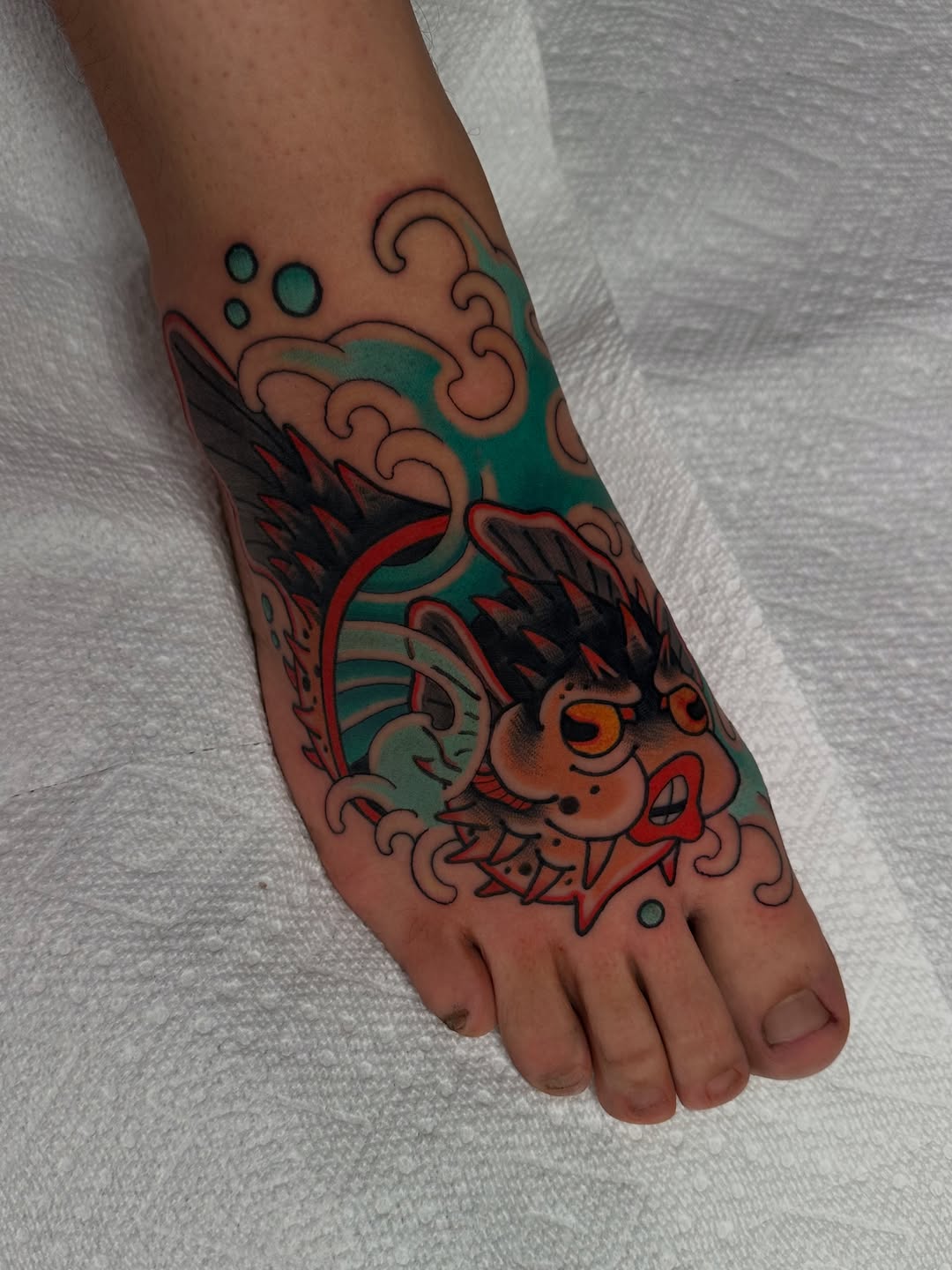 Colorful koi fish tattoo on foot and ankle with orange, teal, and red ink featuring decorative waves and bubbles.