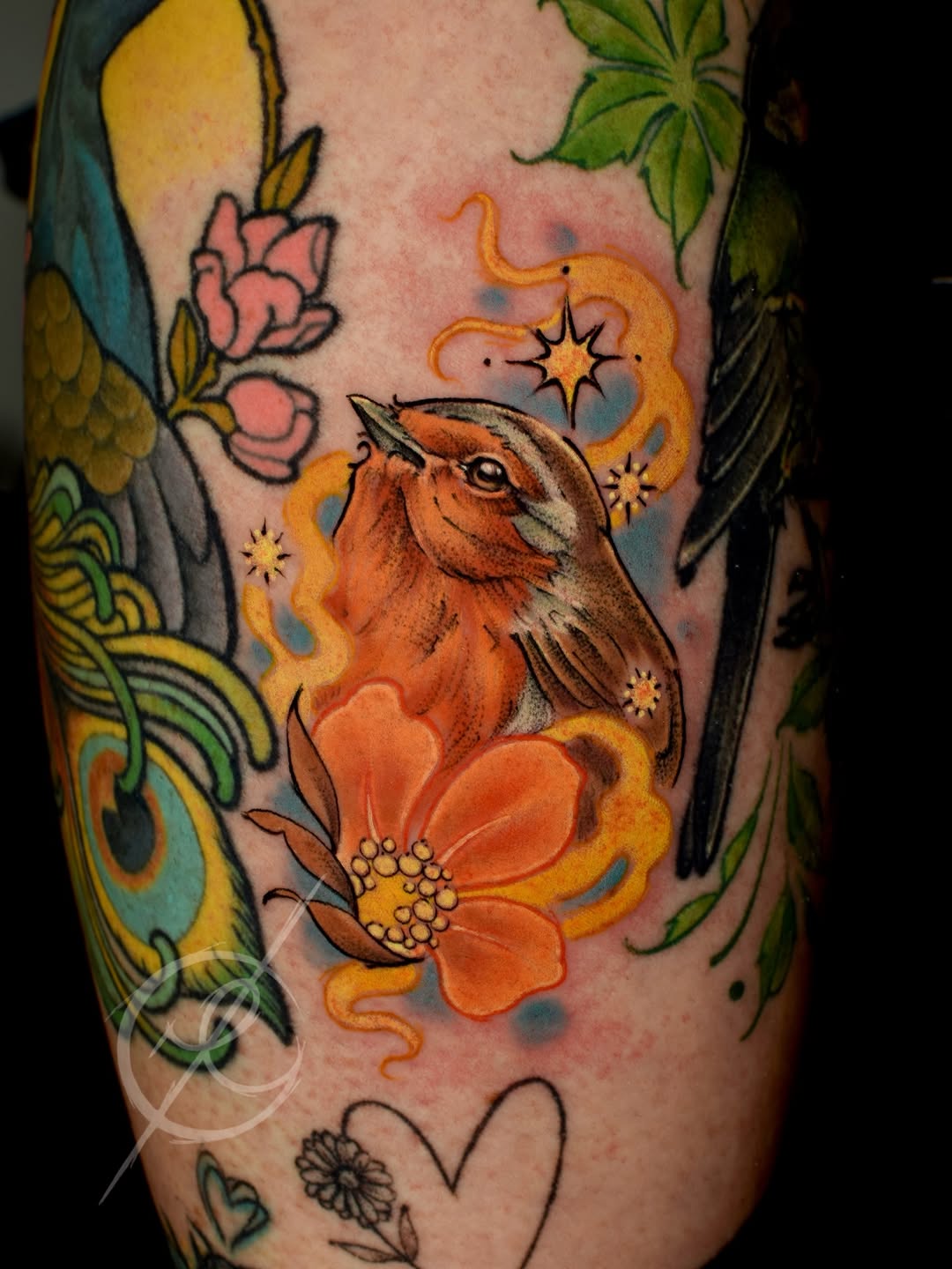 A colorful leg tattoo featuring an orange bird with magical flames and stars, surrounded by flowers and botanical elements in orange, yellow, green, and blue.