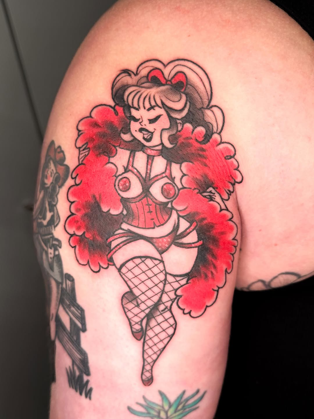 Colorful tattoo on upper arm featuring a pinup-style figure with voluminous red pom-pom details, black line work, and fishnet stockings.