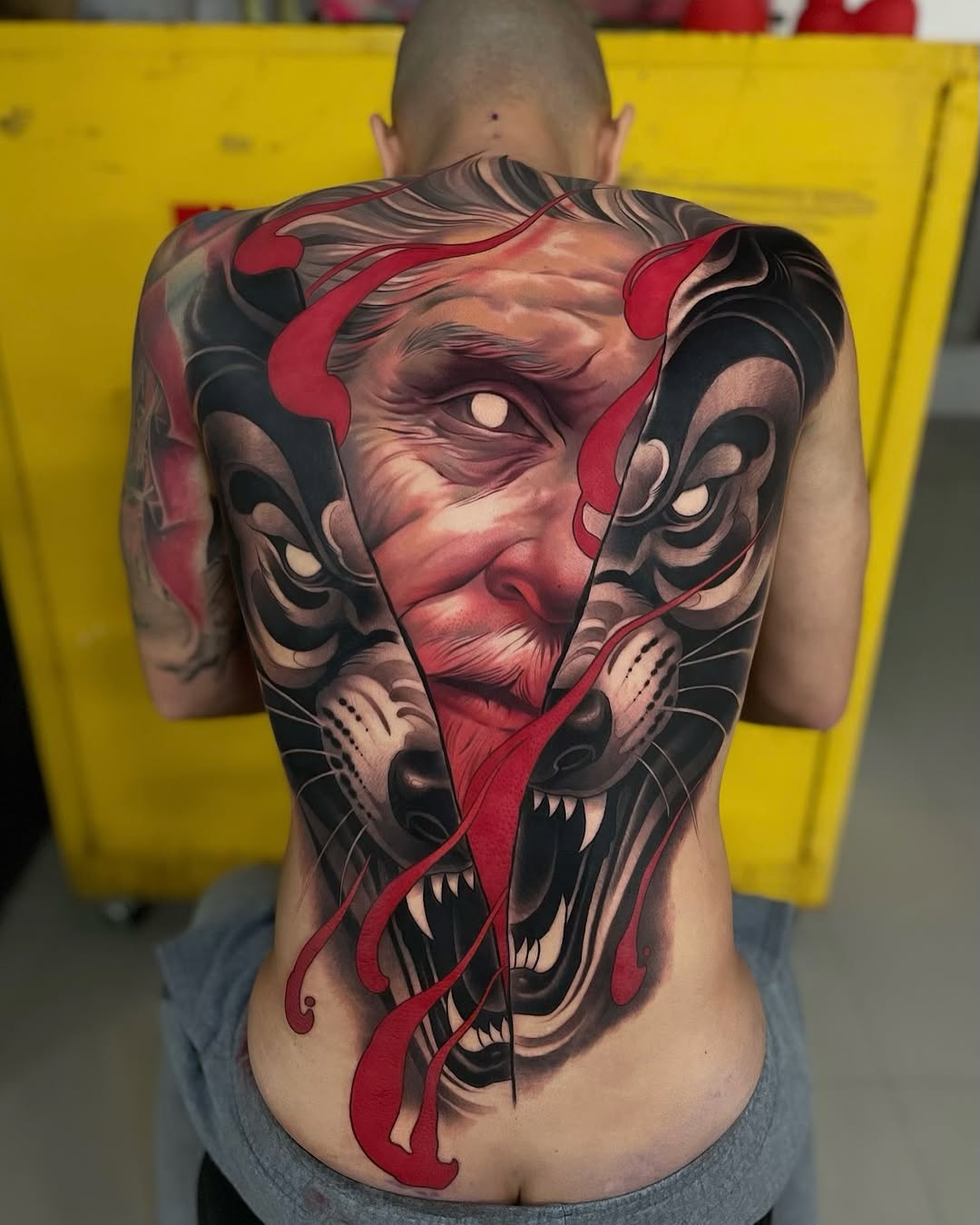 Large back tattoo featuring a demon face with red horns and accents, black details, and red curved elements extending downward on bare skin.