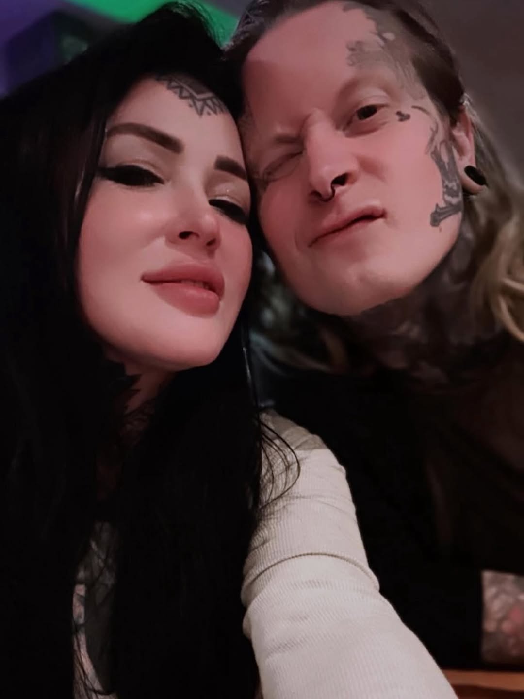 Two people with visible face and neck tattoos pose closely together against a colorful background.