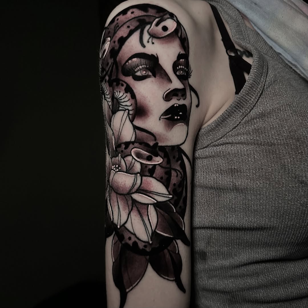 Portrait tattoo on upper arm featuring a woman's face with ornamental patterns, floral elements, and dark shading in black and purple tones.