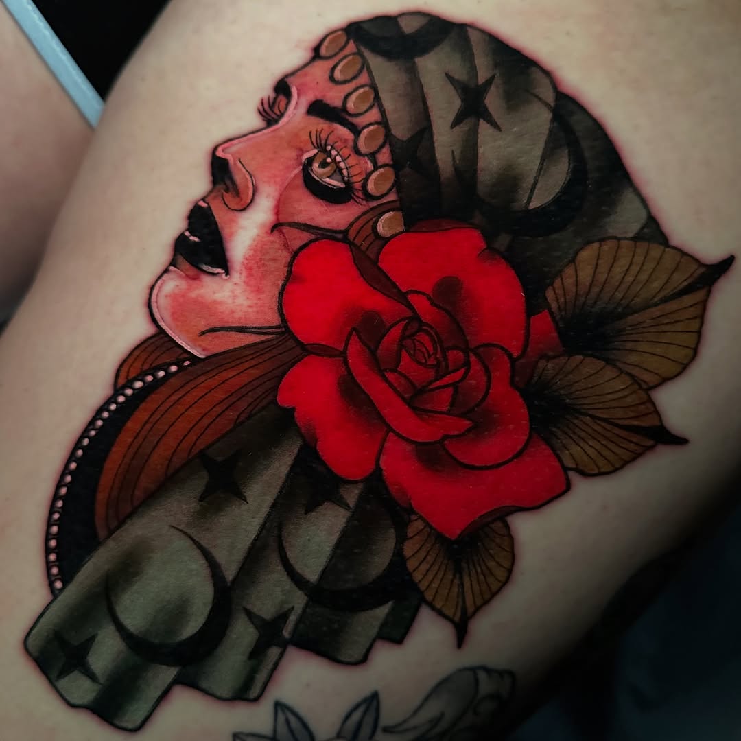 Colorful shoulder tattoo featuring a woman's profile with a large red rose, ornamental wings, and decorative elements in red, pink, brown, and black tones.