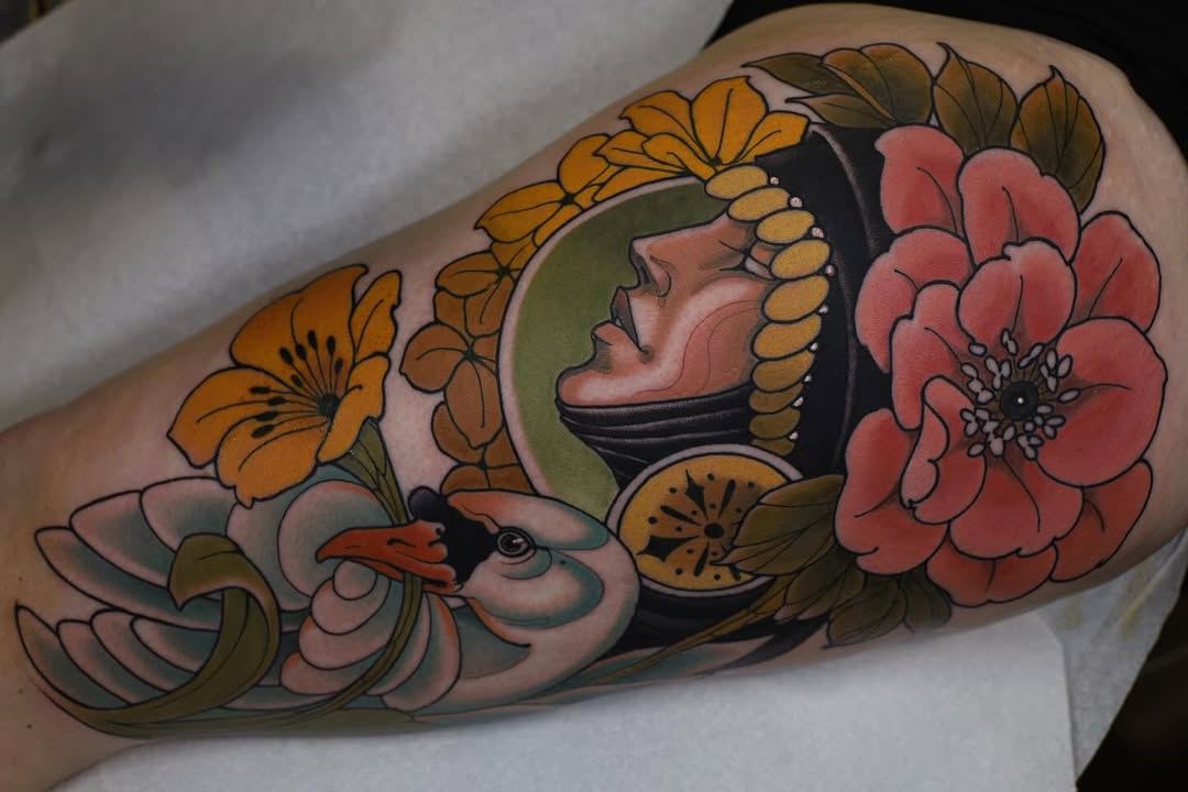 Colorful shoulder tattoo featuring a profile face with golden ornamental headdress, surrounded by orange and red flowers with green leaves and a decorative bird.
