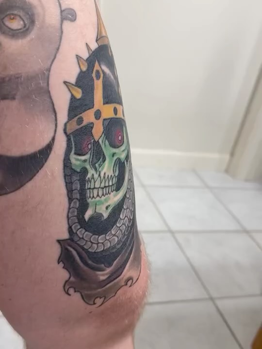 Skull with golden cross and green details tattooed on upper arm, featuring black shading and red eye accents.