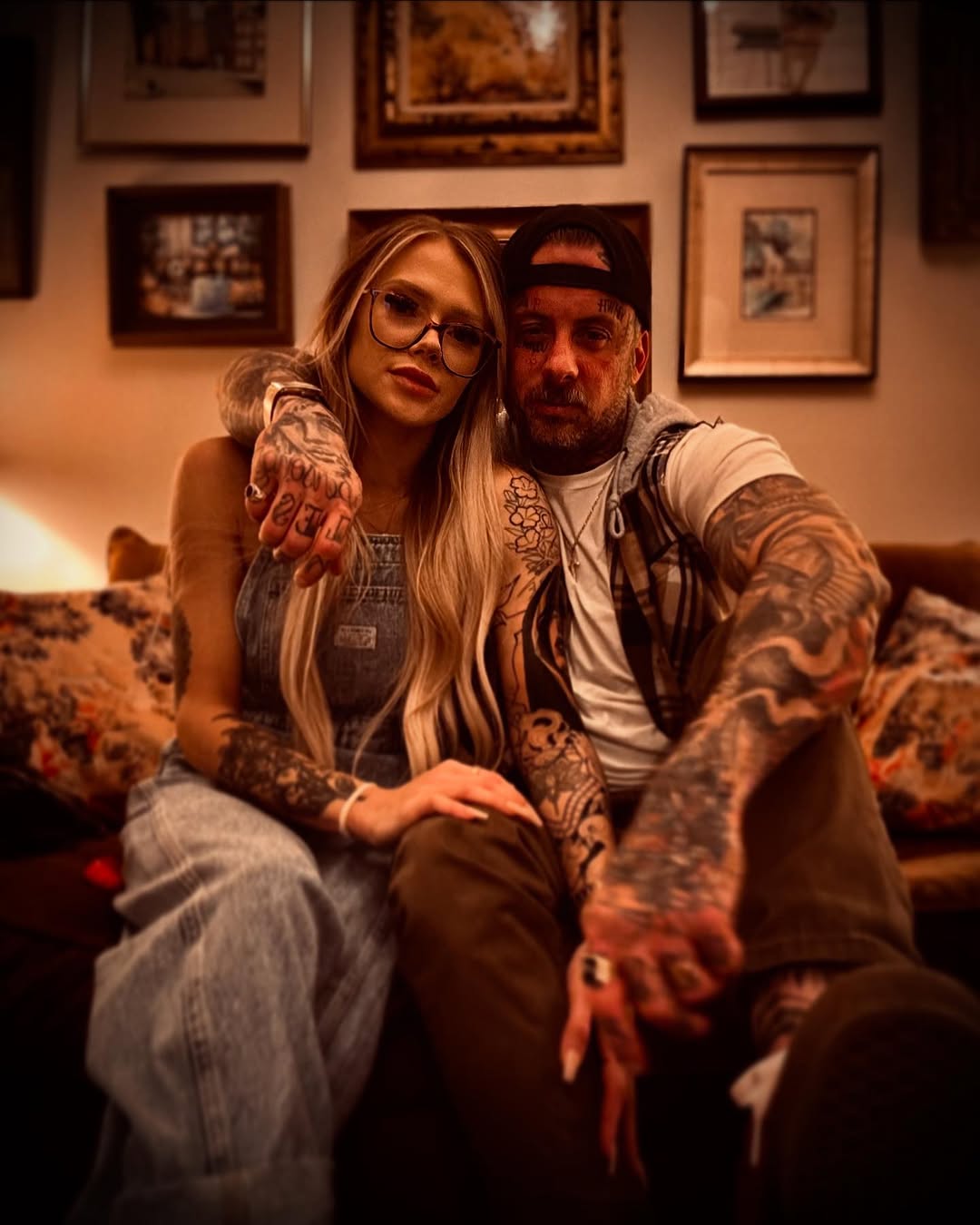 Two tattooed individuals pose together on a couch against a wall of framed photographs, displaying extensive arm and hand tattoos in dark ink.