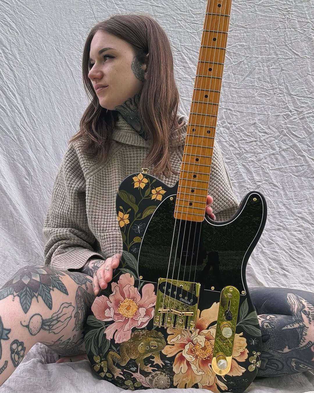 Woman with full arm tattoos featuring flowers and botanical elements in black, pink, and gold displays a custom electric guitar with matching floral designs.