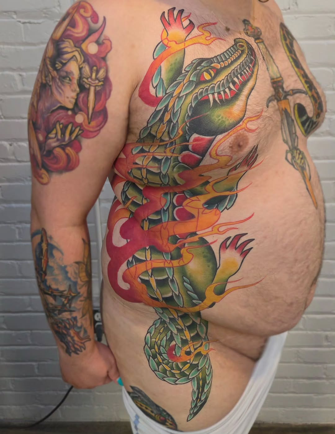 Colorful dragon tattoo covering the torso and extending down the leg, featuring red, green, yellow, and orange hues with detailed scales and flames.