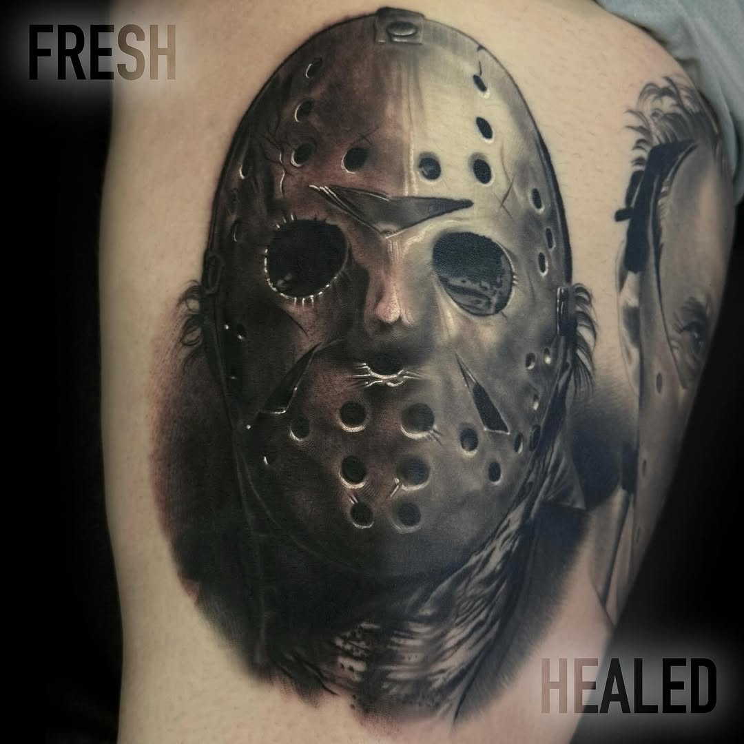 Jason Voorhees hockey mask tattoo on upper arm showing the masked face with lacing details and feather elements in grayscale and black ink.