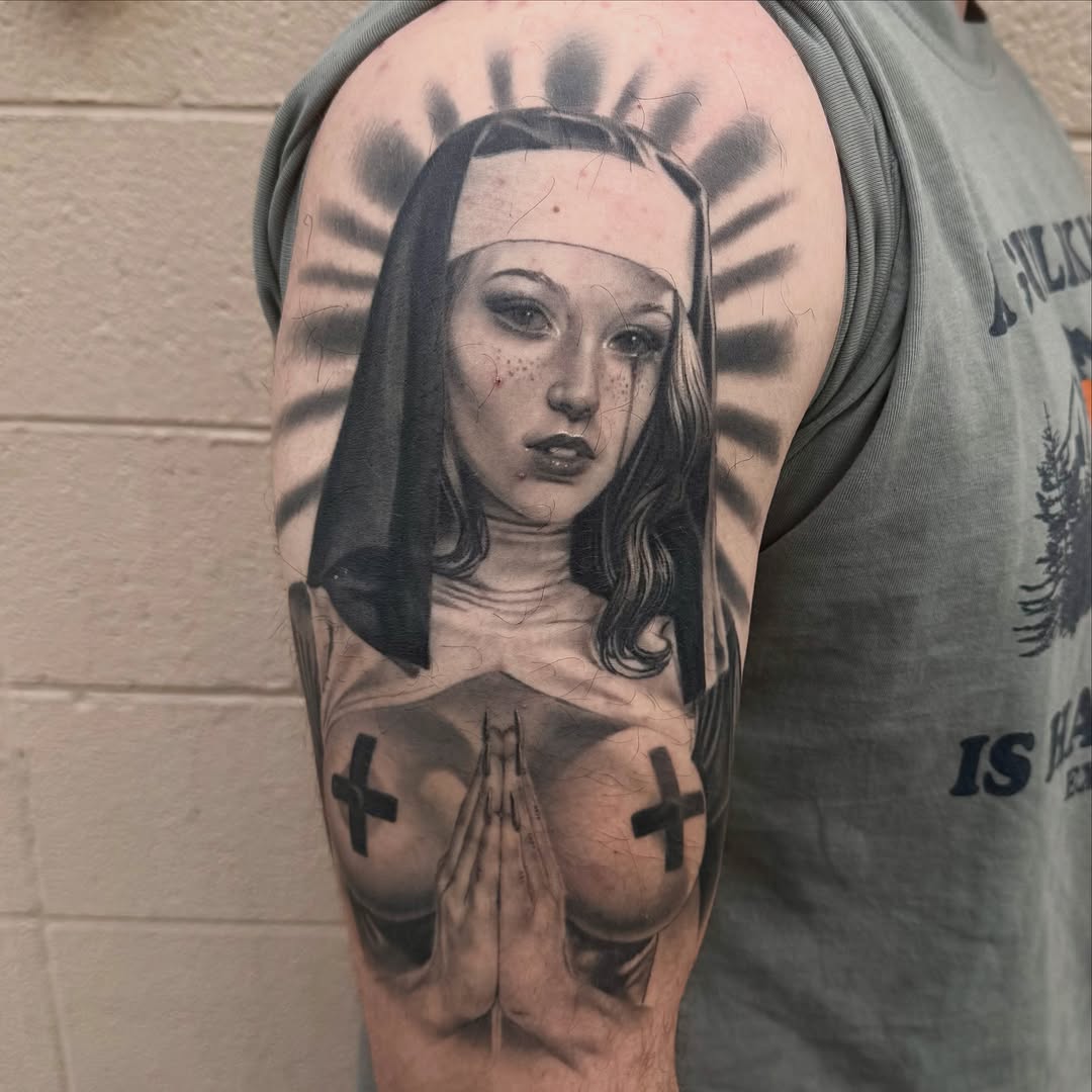 Portrait tattoo on upper arm of a woman with long hair, headpiece, halo, crosses, and praying hands in black and grey ink.