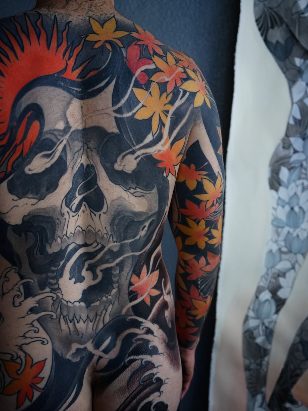 Large skull with red and gold flowers on dark background covering person's back and shoulder, with ornamental blue swirling details.