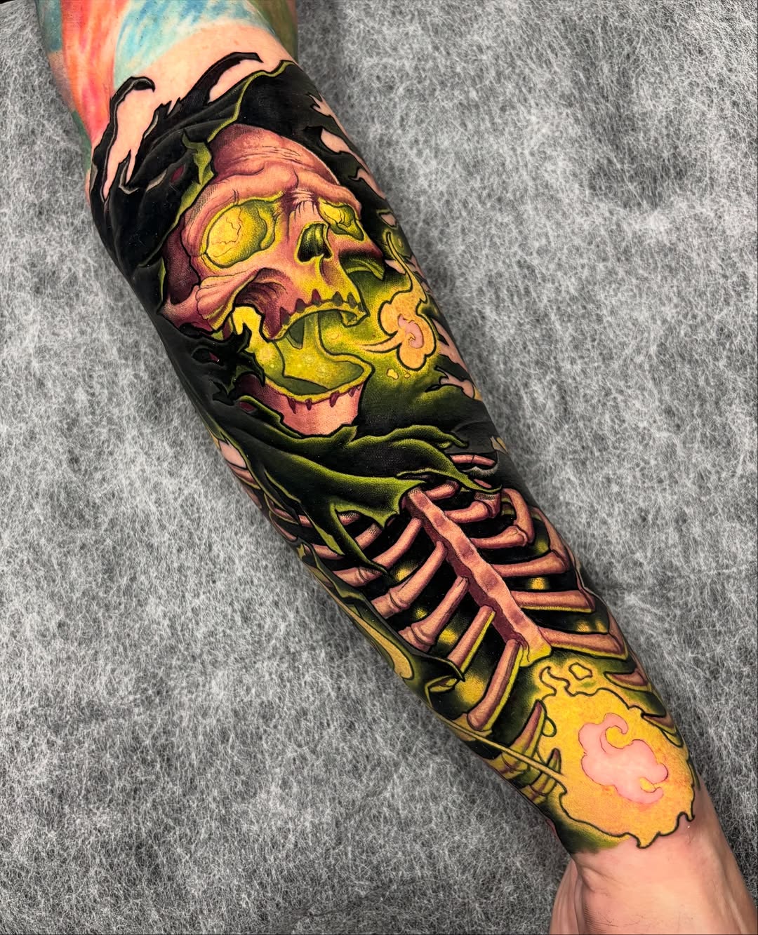 Colorful skull and skeleton ribcage tattoo on forearm featuring yellow, green, pink, and black ink with decorative flames at the wrist.