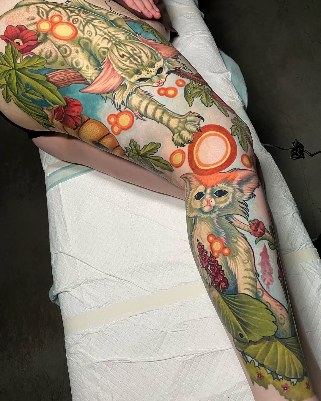 Full leg sleeve tattoo featuring two cats with flowers, berries, botanical elements, and circular orange motifs on a person's thigh and calf.
