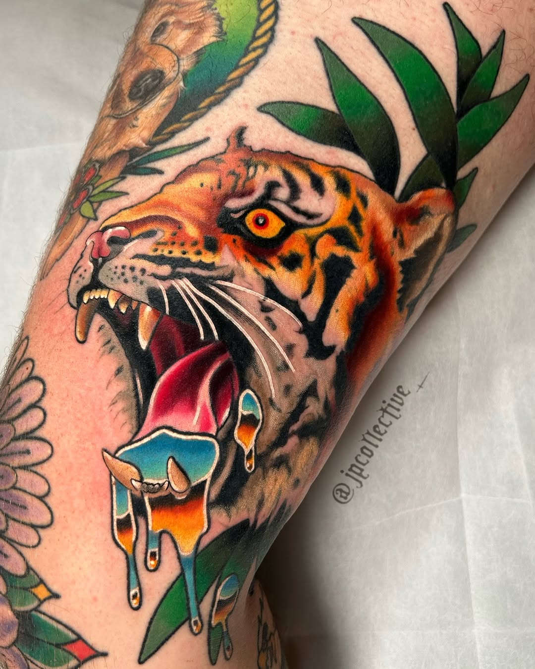 Colorful tiger head tattoo on an arm with open mouth, orange and red tones, surrounded by green foliage and dripping paint effects.