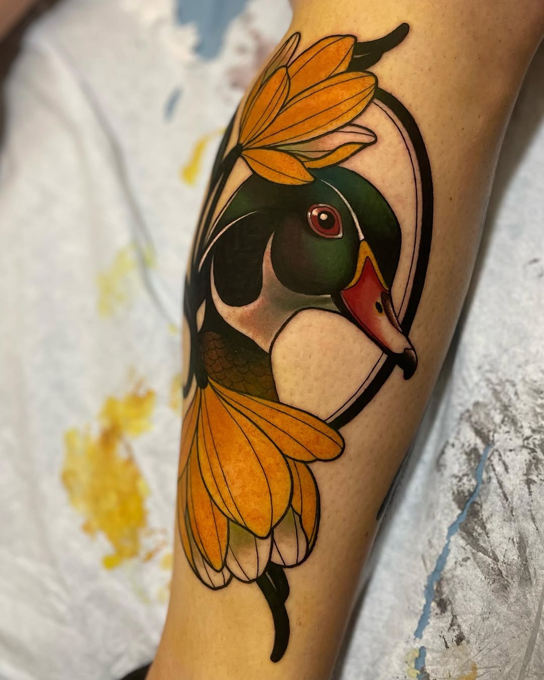 Colorful tattoo of a duck's head with orange and yellow wing feathers framing it, featuring green plumage, red markings, and a red eye on the inner forearm.