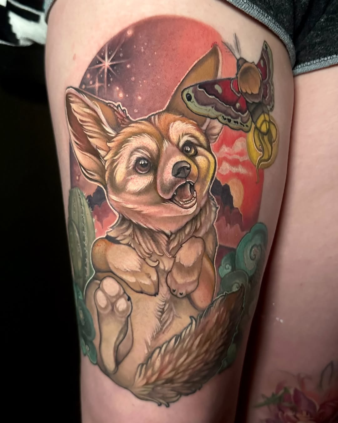 Colorful thigh tattoo featuring a happy corgi with butterflies and moths against a red moon and green foliage background.