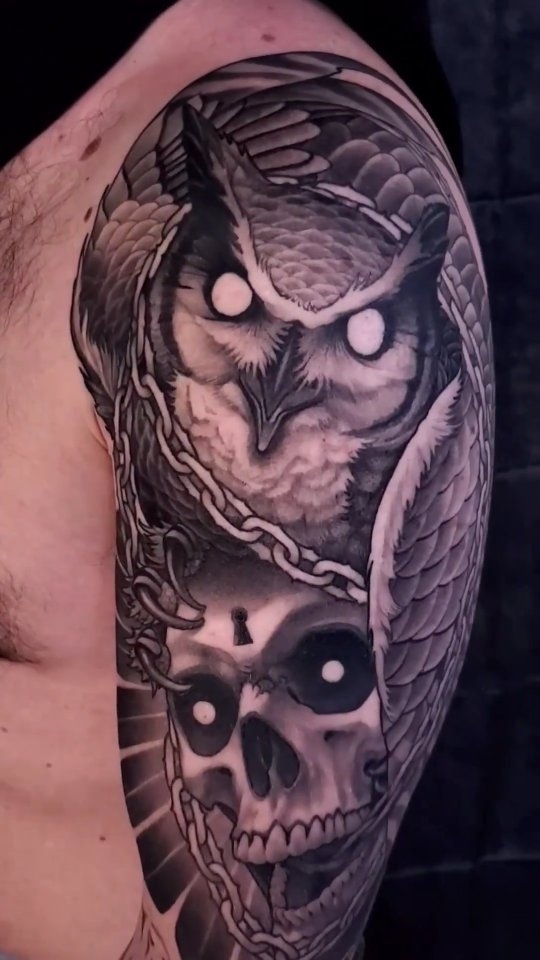 Owl with spread wings perched above a skull, both with glowing eyes, tattooed on upper arm in shades of gray and black.