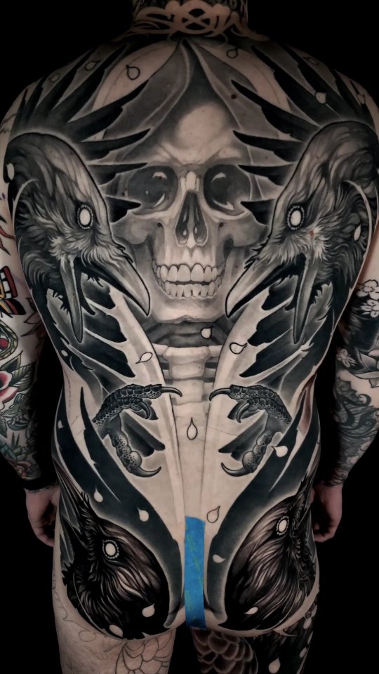Large-scale back tattoo featuring a central skull with ornamental crown, flanked by birds and snakes in grayscale with blue accent stripe.
