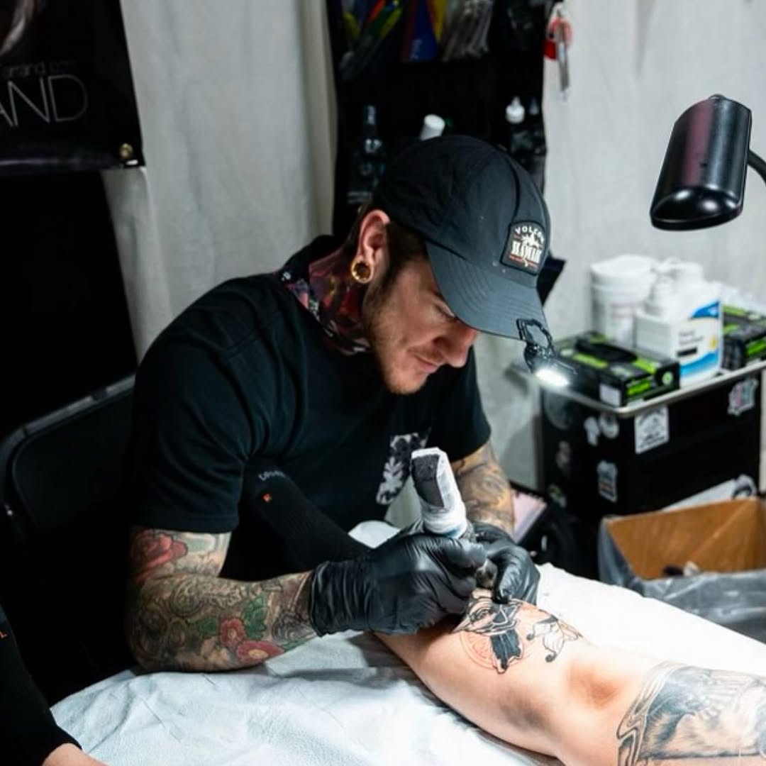 Tattoo artist wearing black gloves and cap applies a colorful floral design to a client's forearm in a studio setting.