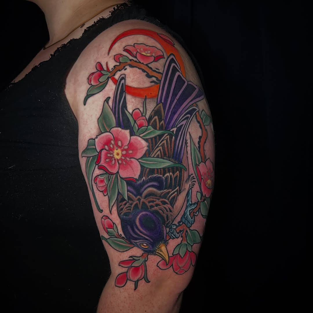 Colorful upper arm tattoo featuring birds with spread wings, pink flowers, red ribbons, and green leaves in red, purple, blue, and orange tones.