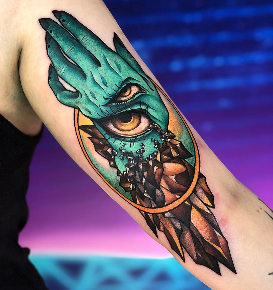Dragon eye tattoo on upper arm featuring turquoise scales, golden iris, and geometric shattered pattern below in orange and brown tones.